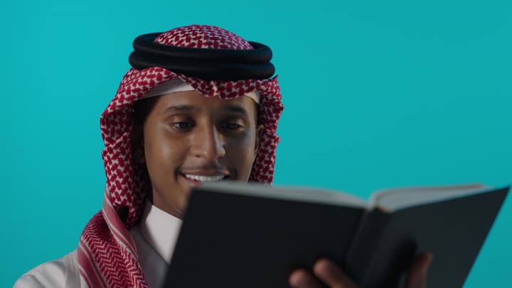 شاب سعودي بالزي التقليدي يقرأ كتاباً في استوديو خلفية زرقاء