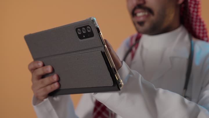 تصفح مواقع التواصل الاجتماعي، إدارة شؤون الأعمال وتنظيمها عبر الاجهزة التقنية، دمج العمل مع التقنية والتكنولوجيا، برجل عربي خليجي سعودي يرتدي الزي التقليدي والغترة يعمل على جهاز التابلت المحمول ، خلفية ملونة