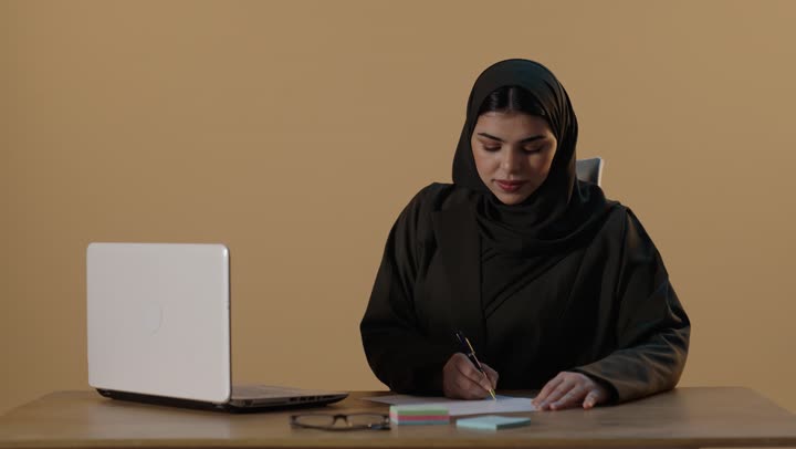Saudi Businesswoman Writing at Desk with Laptop