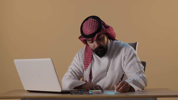 Saudi Businessman Working on Laptop and Writing in Office