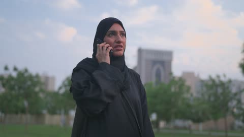 امرأة عربية بالعباءة تستخدم الهاتف في حديقة المدينة مساءً